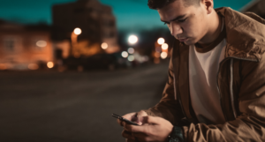A teenager looking at their smart phone on a street at night time.